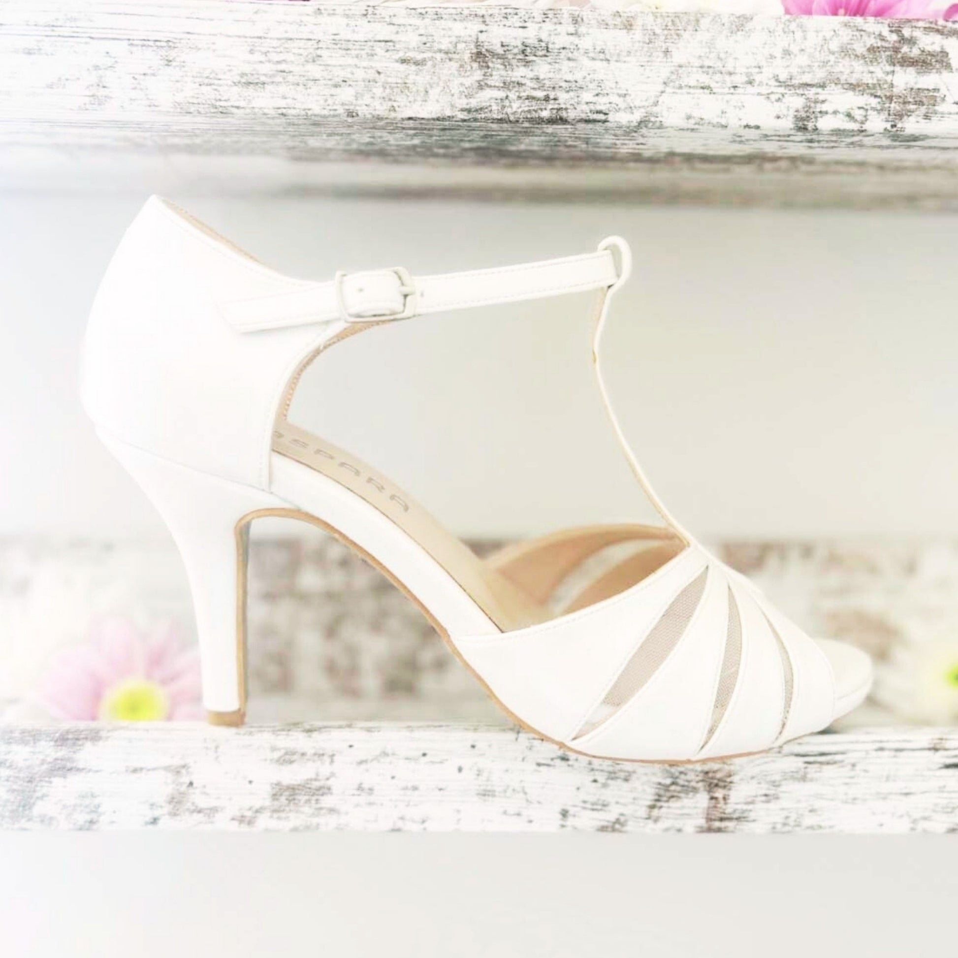Elegant ivory wedding shoes resting on a shelf surrounded by vibrant flowers.